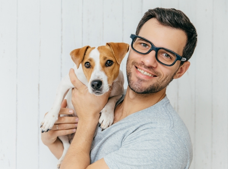 A man with dark hair and glasses holds a small brown and white dog in front of a white wooden wall.
